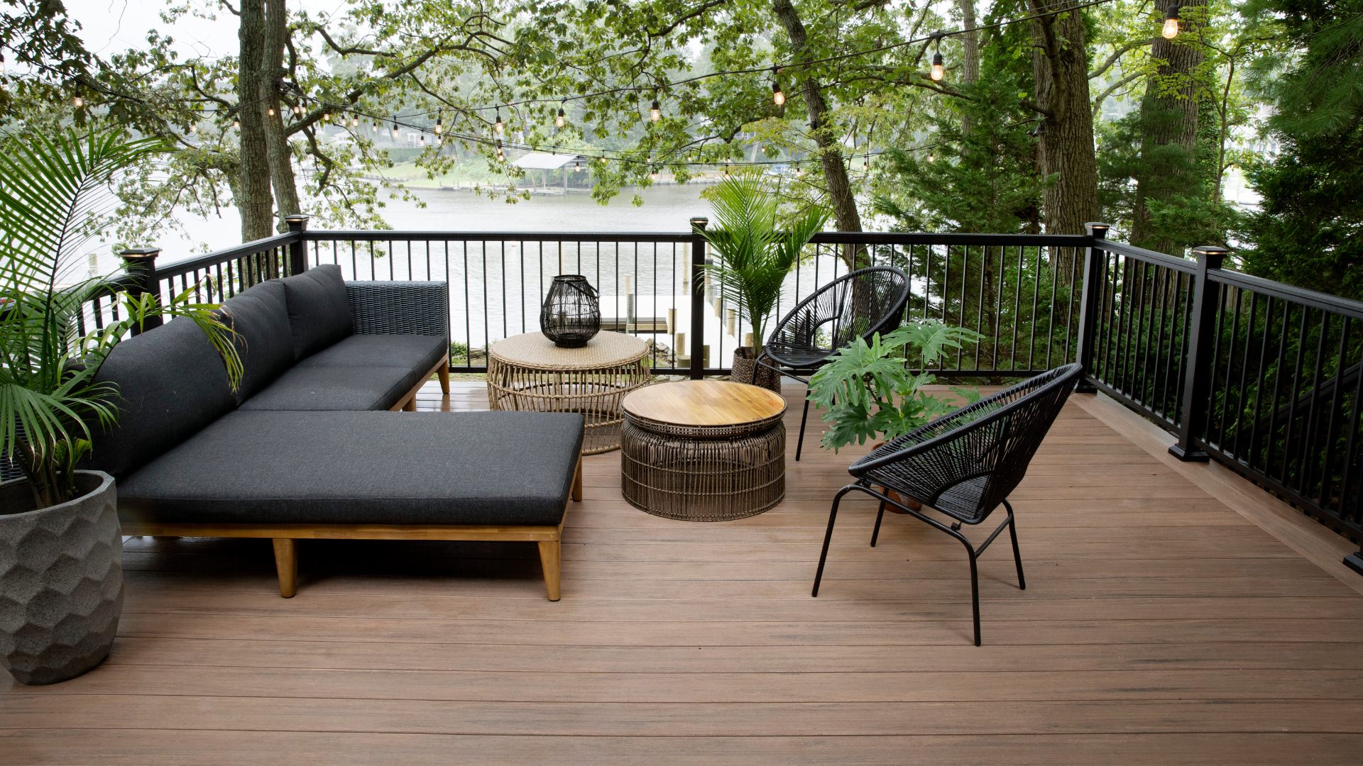 Mahogany color Timbertech deck with modern couches and party lights overlooking a lake in the afternoon. This image is promoting custom deck design by Deck Teck