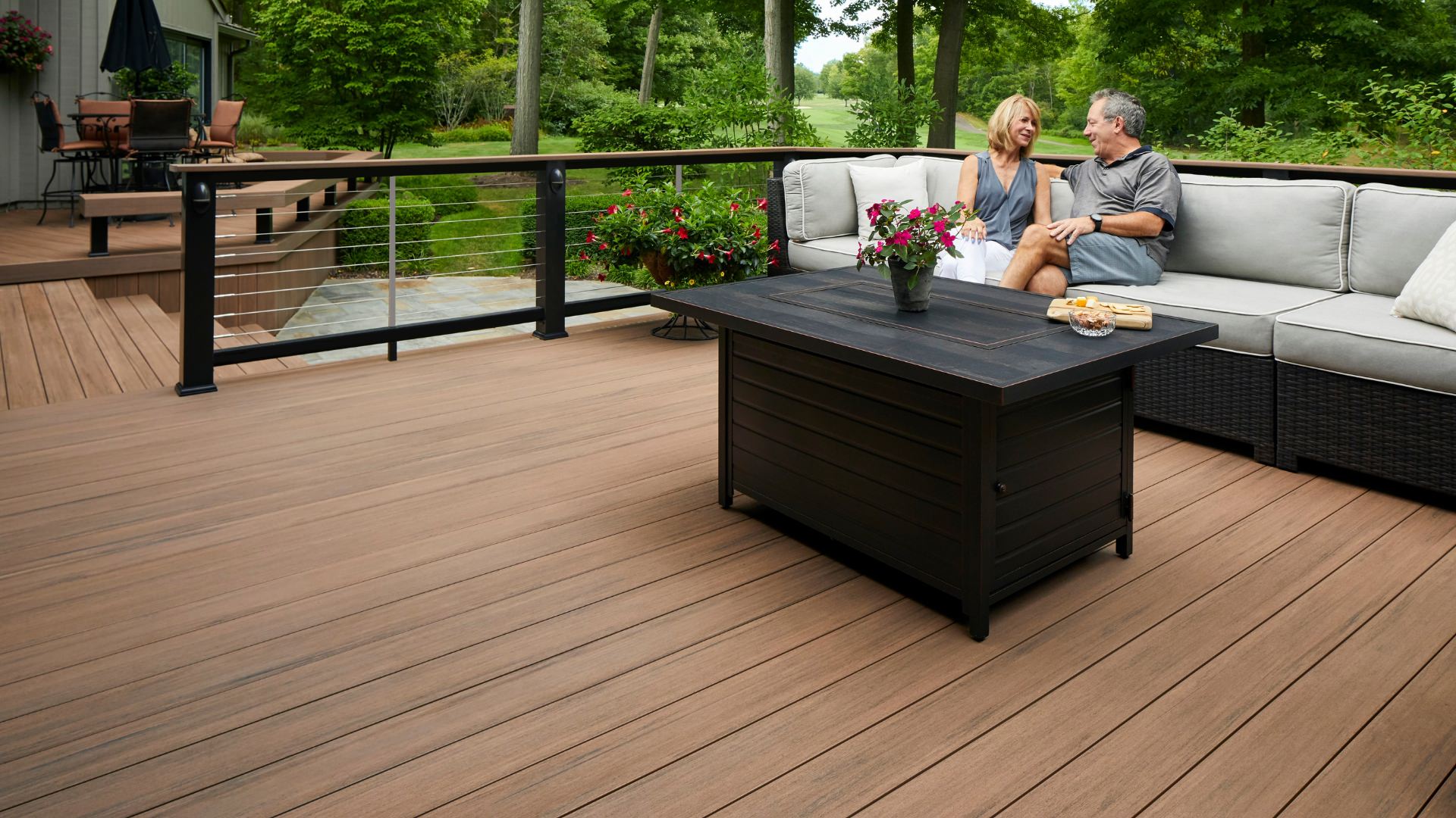 Mahogany color Timbertech deck with modern couches and a couple in their 50's looking at each other enjoying the couch. This image is promoting custom deck design by Deck Teck