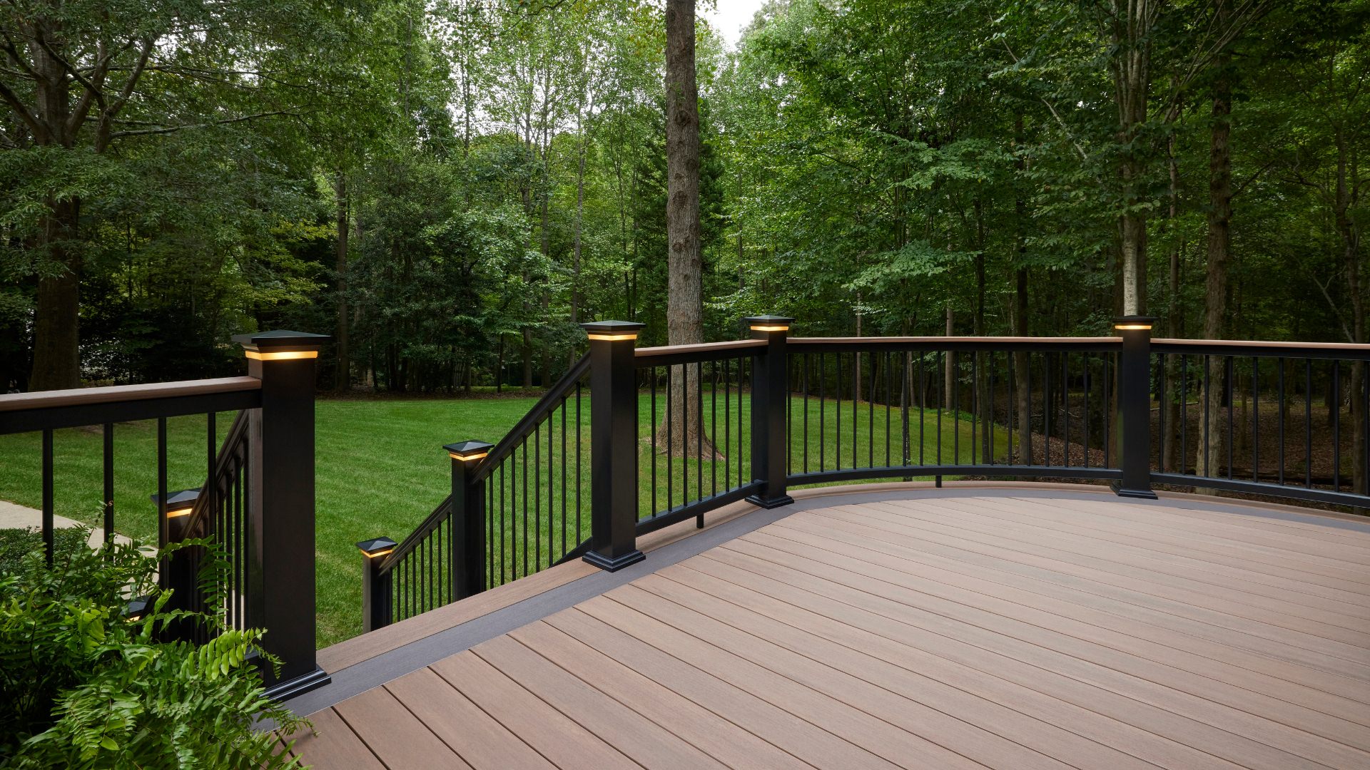 Mahogany color Timbertech deck with grey inlay and black railing overlooking a freshly mowed grass backyard in the woods. This image is promoting custom deck design by Deck Teck